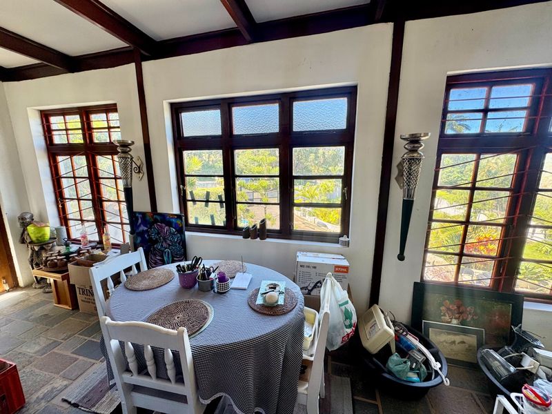 A sunlit living area featuring exposed timber ceiling beams, stone tile flooring, and large timber-framed windows with security bars.