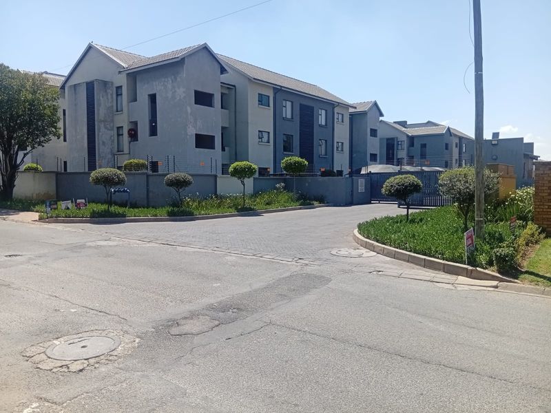 Street view of an apartment complex featuring a paved entrance, perimeter wall with electric fencing, and multi-story residential buildings with tiled roofs.