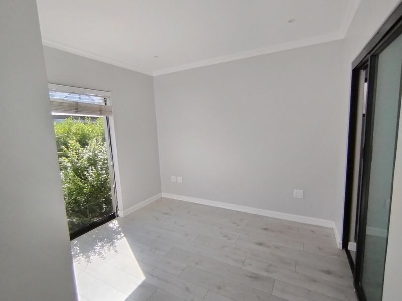 A bright, empty bedroom featuring modern light-toned laminate flooring, recessed ceiling lighting, and a sliding glass door providing natural light and garden views.