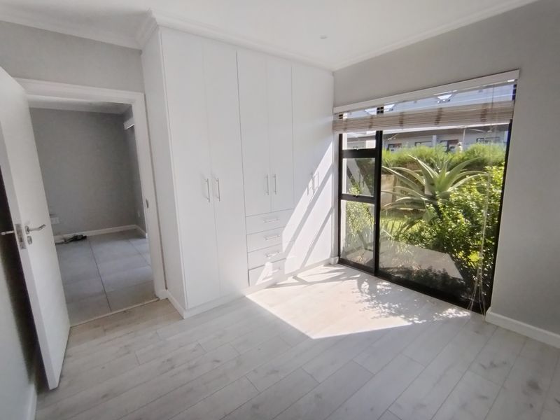 A bright, empty bedroom featuring modern white built-in cupboards, light-toned laminate flooring, and a large window overlooking the garden.