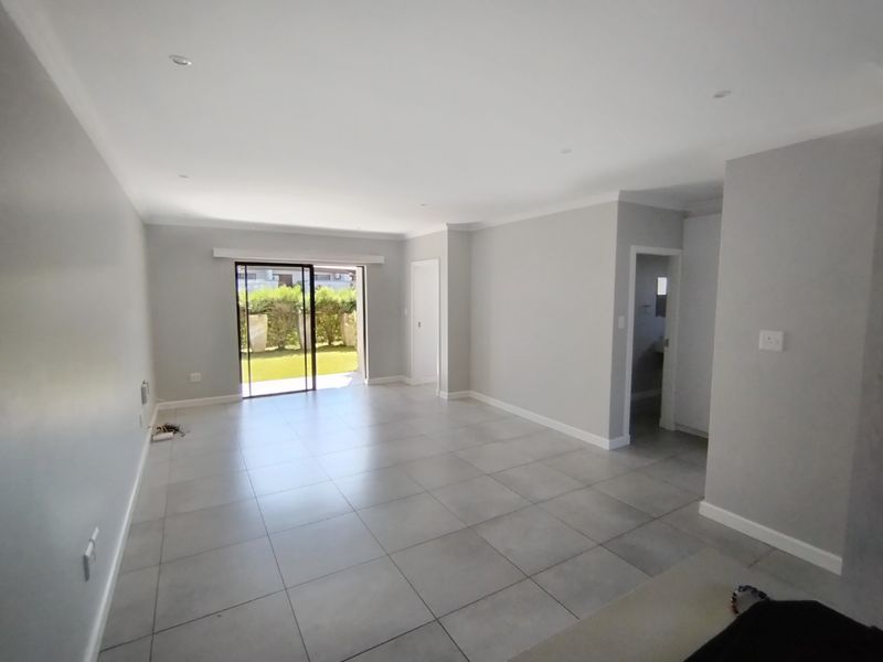 A spacious, empty living area featuring neutral grey walls, large format floor tiles, and a sliding glass door providing access to the outdoor space.