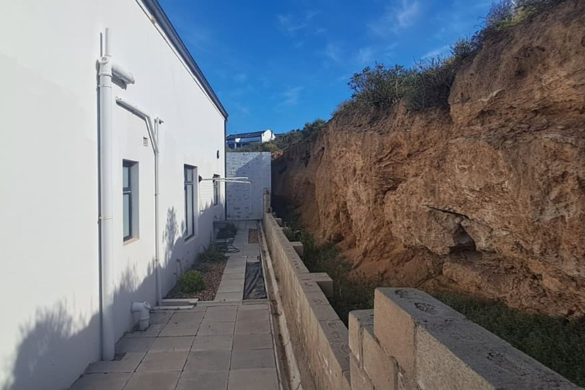 A narrow rear exterior walkway featuring a paved path alongside the building, bordered by a natural rock face and an unfinished retaining wall.