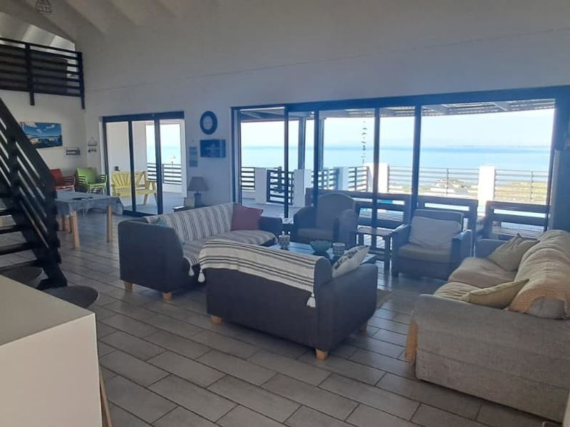 Spacious open-plan living area featuring a built-in fireplace, staircase to a mezzanine level, and large sliding doors opening to a scenic coastal view.