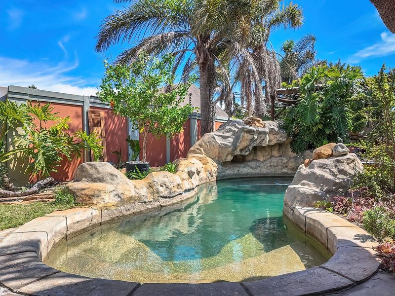 A landscaped outdoor pool area featuring a naturalistic rock-style swimming pool, established garden, and perimeter boundary wall.