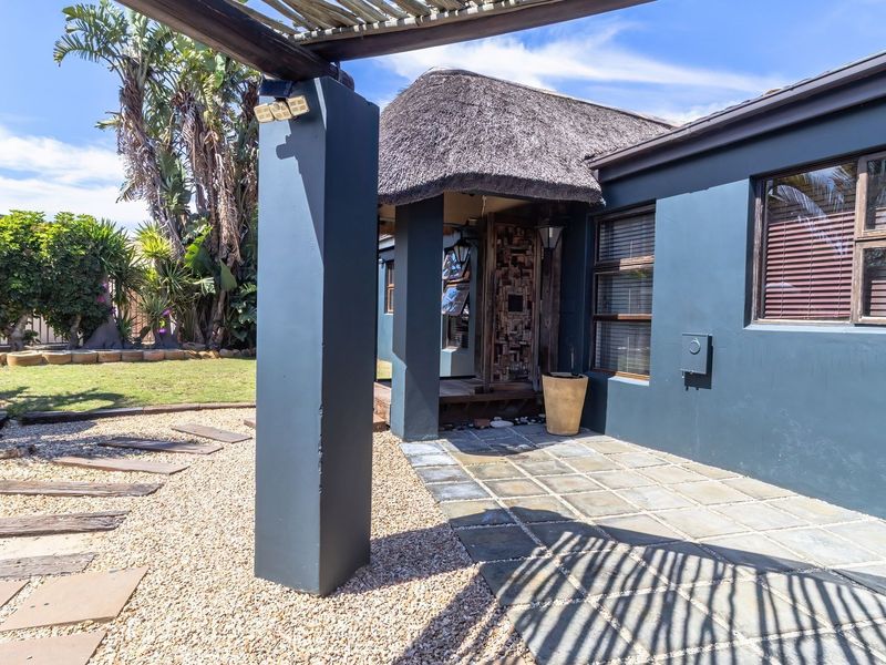 Rear exterior view featuring a thatched roof, paved patio area, and timber pergola, set within a landscaped garden with gravel pathways.