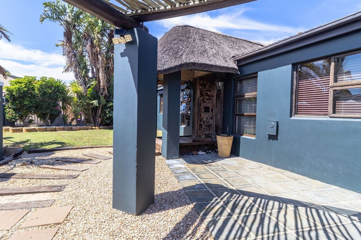 Rear exterior view featuring a thatched roof, paved patio area, and timber pergola, set within a landscaped garden with gravel pathways.