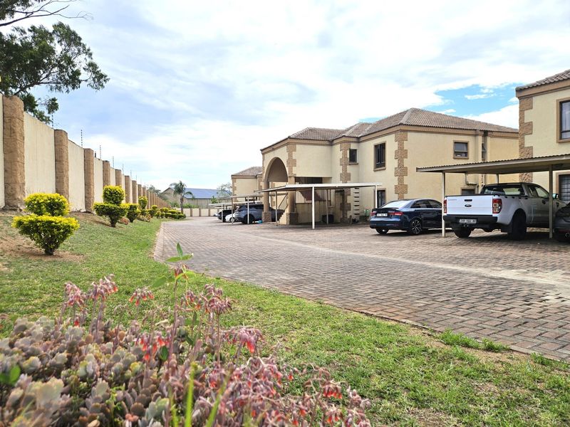 Exterior view of an apartment complex featuring a paved driveway, covered carports, and a secure perimeter wall with electric fencing.