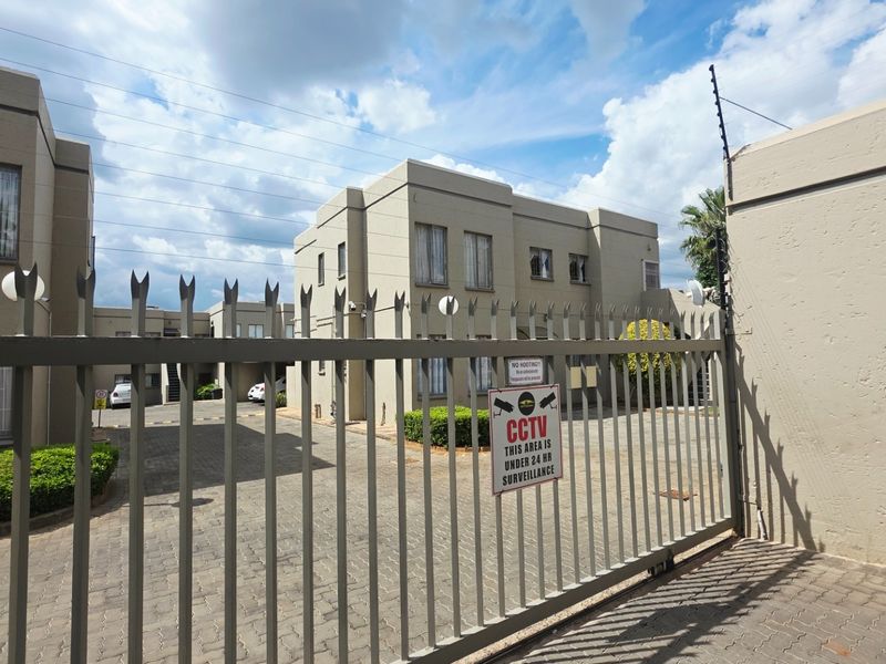 Front entrance view of a townhouse complex featuring a secure metal gate, paved driveway, and perimeter electric fencing.