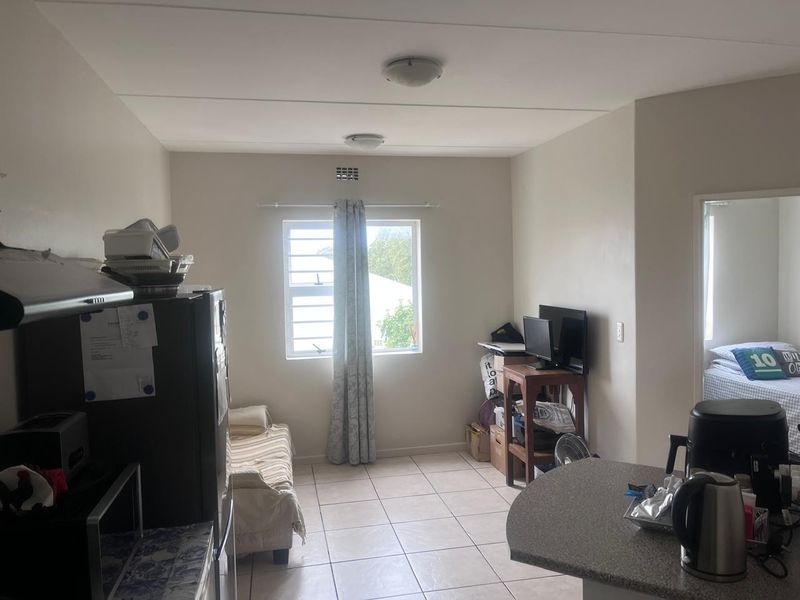 An open-plan living area featuring tiled flooring, a breakfast counter, and a window with security bars, providing a functional and versatile living space.