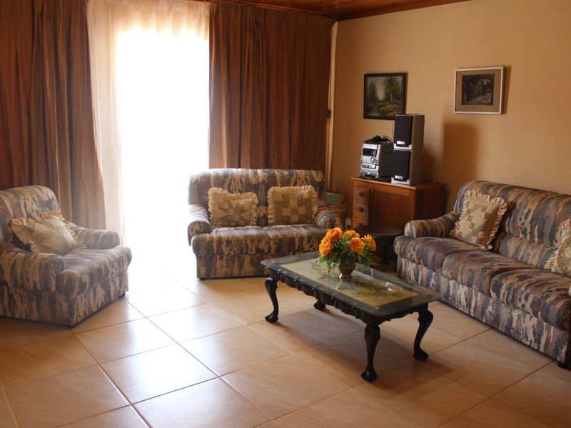A living room featuring tiled flooring and timber ceiling trim, with natural light entering through a large window covered by curtains.