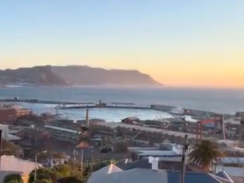 Elevated panoramic view overlooking the harbour, industrial port facilities, and distant coastal mountains at sunset.