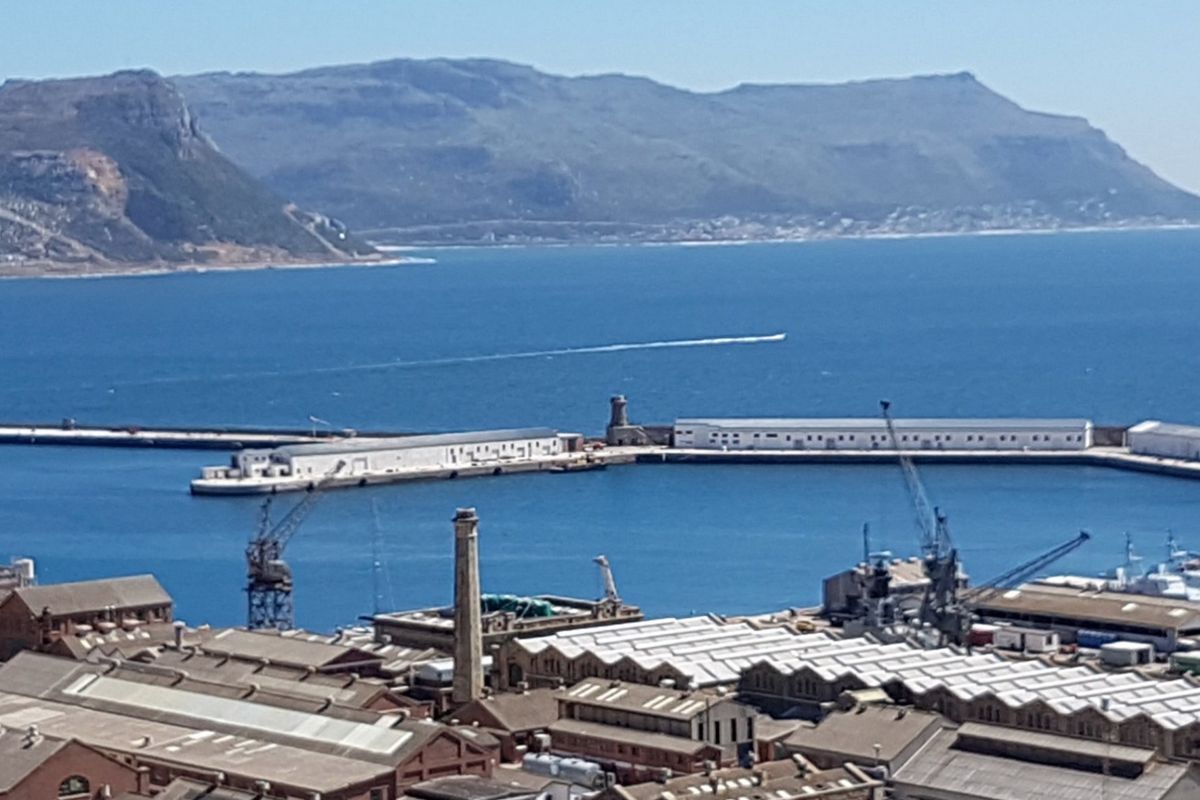 An elevated view overlooking a commercial harbour, industrial buildings, and the surrounding coastline with mountain scenery in the background.