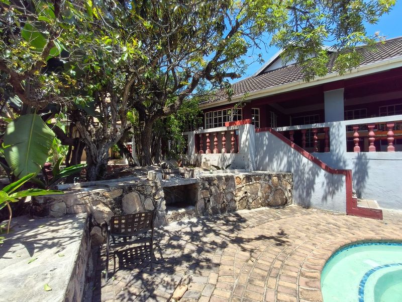 Outdoor pool area featuring a stone-clad built-in braai, brick paving, and a raised patio with concrete balustrades under mature trees.