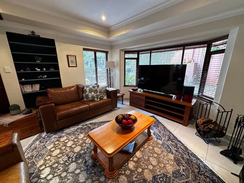 A comfortable living room featuring a tray ceiling, tiled flooring, built-in shelving, and a large bay window with venetian blinds.
