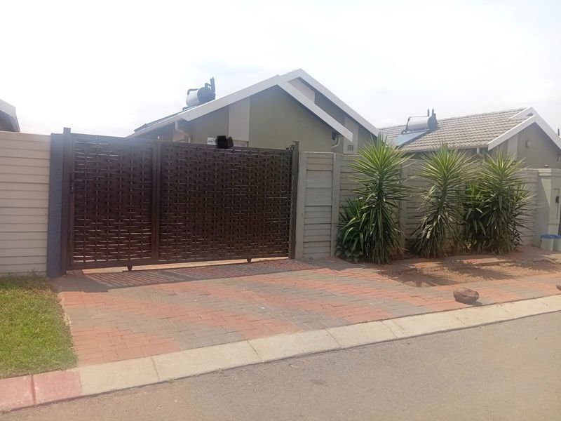 Front view of the property featuring a secure metal driveway gate, paved driveway, and precast boundary wall with an installed solar water heater.