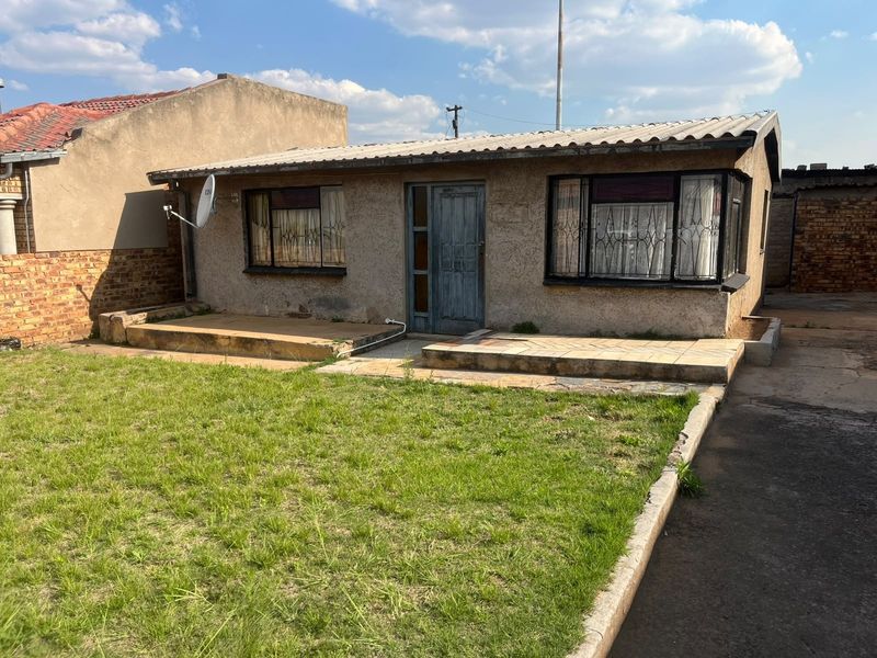 Rear view of a modest house featuring a small tiled patio, grass lawn, and brick boundary walls, showing signs of age and weathering.