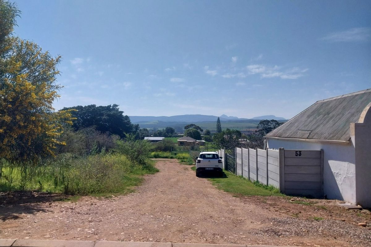 Street view of a residential property featuring a dirt driveway, precast concrete boundary wall, and distant mountain views.