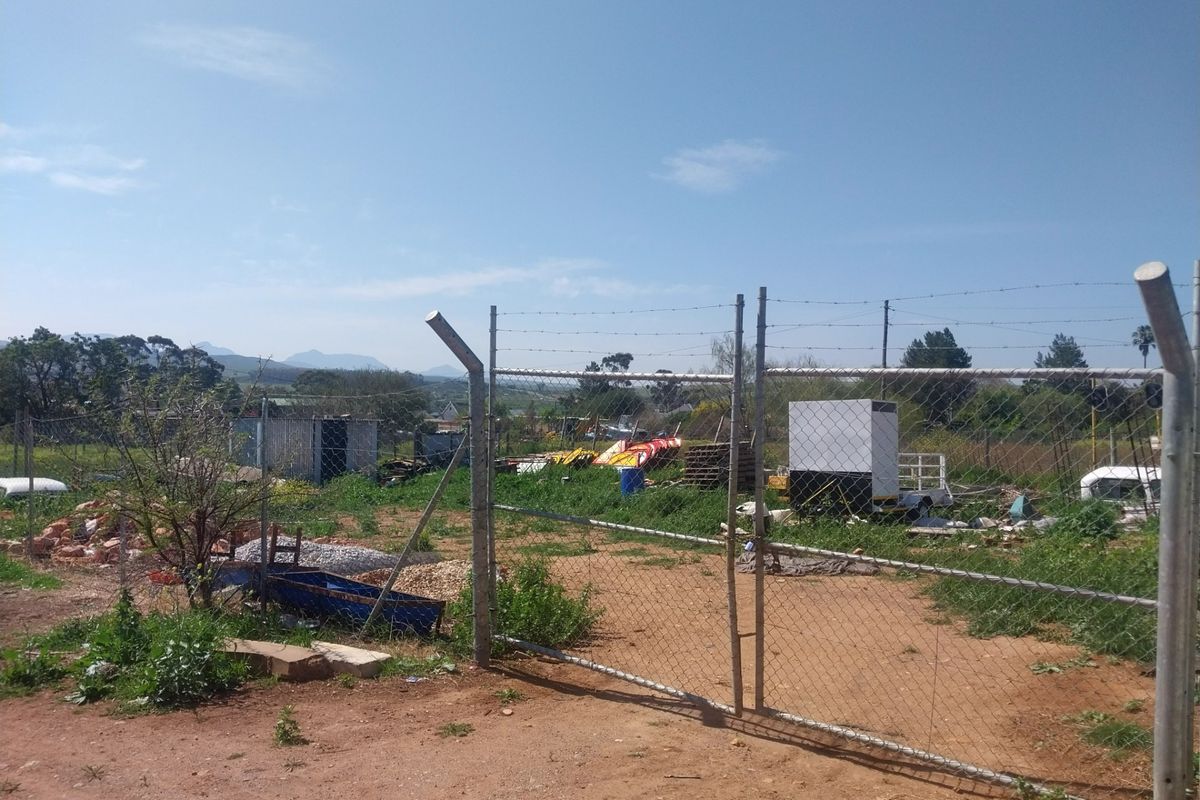 A view of a vacant residential plot secured with chain-link fencing and a metal gate, showing natural terrain and some site debris.