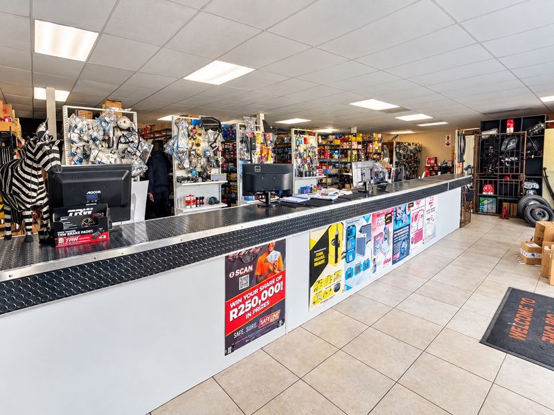 Commercial retail space featuring a long service counter, tiled flooring, suspended ceiling with recessed lighting, and extensive metal shelving for inventory storage.