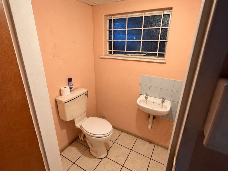 A compact, dated bathroom featuring a toilet, wall-mounted basin, tiled flooring, and a window with security bars.