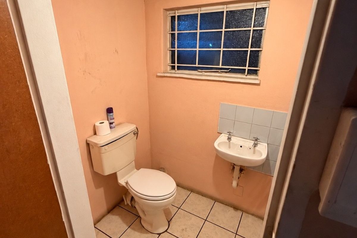 A compact, dated bathroom featuring a toilet, wall-mounted basin, tiled flooring, and a window with security bars.