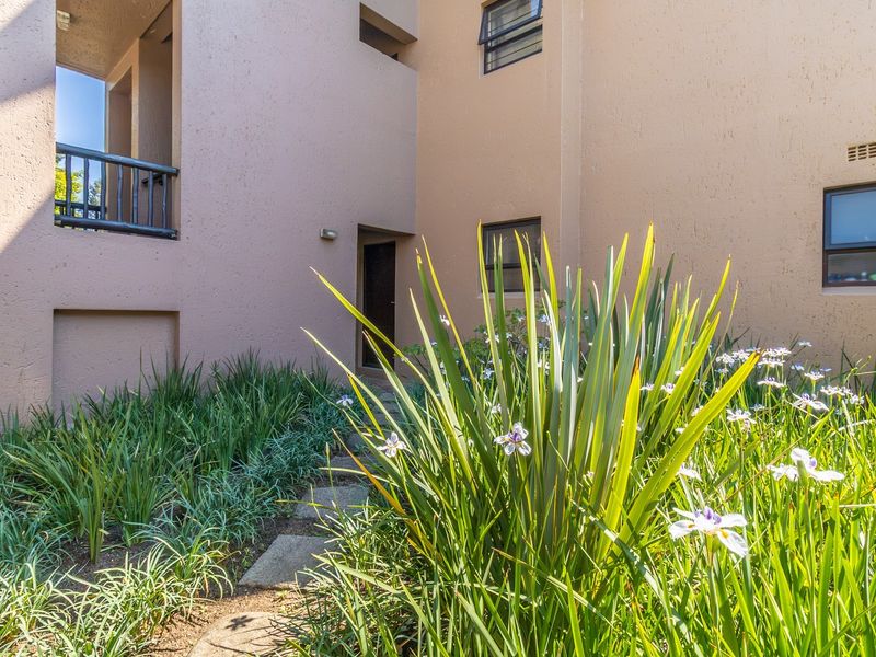 A view of the apartment building's rear exterior, featuring a landscaped garden, stone pathway, and a private balcony with metal railings.