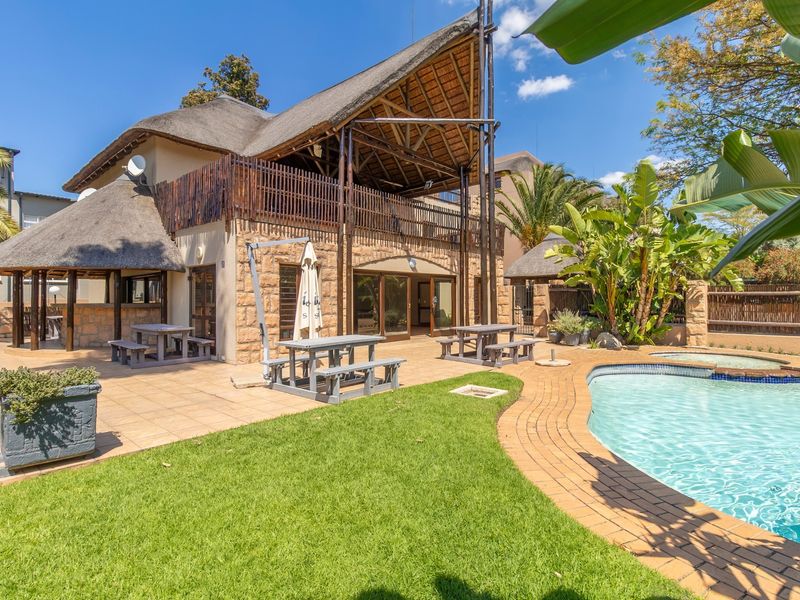A well-maintained outdoor pool area featuring a thatched-roof structure, stone-clad exterior, paved patio, and landscaped garden, ideal for leisure and entertainment.