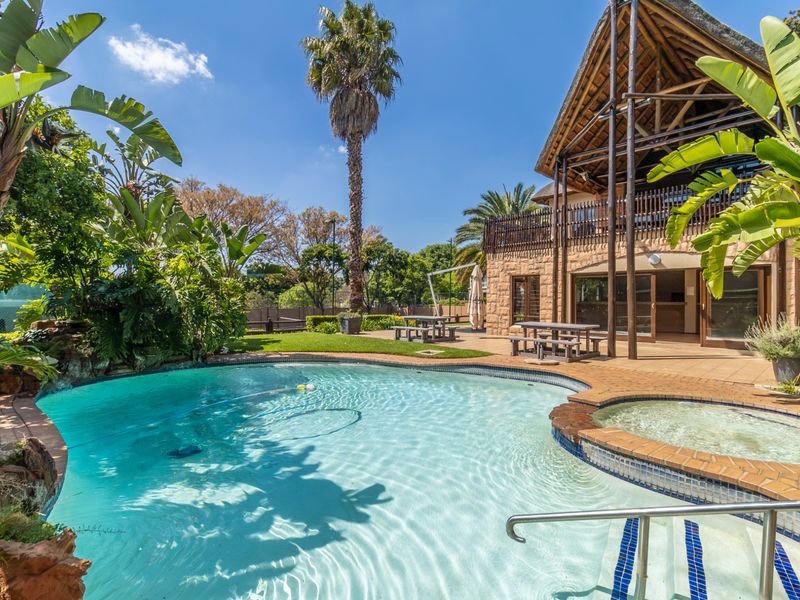 A well-maintained outdoor pool area featuring an integrated spa, stone-clad building exterior, thatched roof structure, and a spacious tiled patio.
