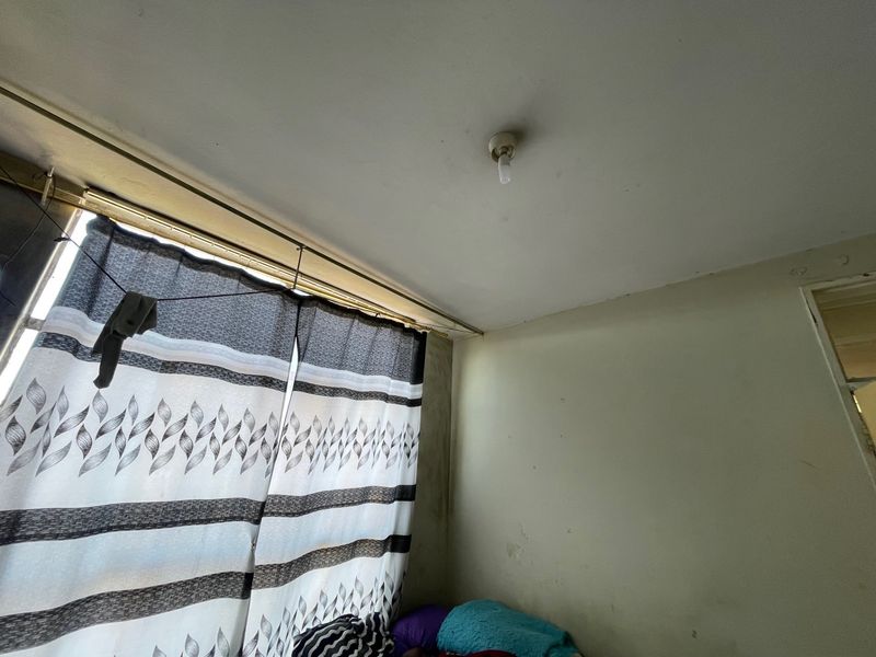 A bedroom interior showing signs of wear on the walls and ceiling, featuring a basic curtain rail and standard light fixture.