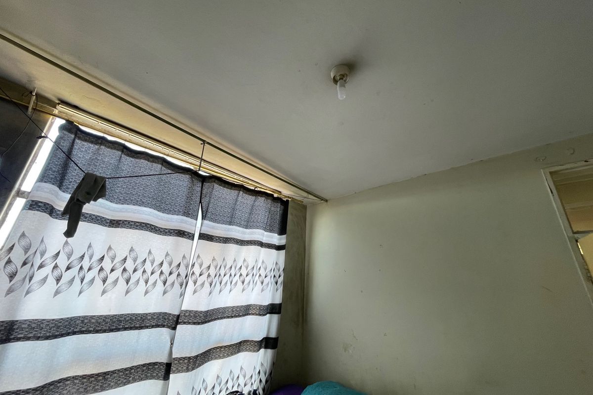A bedroom interior showing signs of wear on the walls and ceiling, featuring a basic curtain rail and standard light fixture.