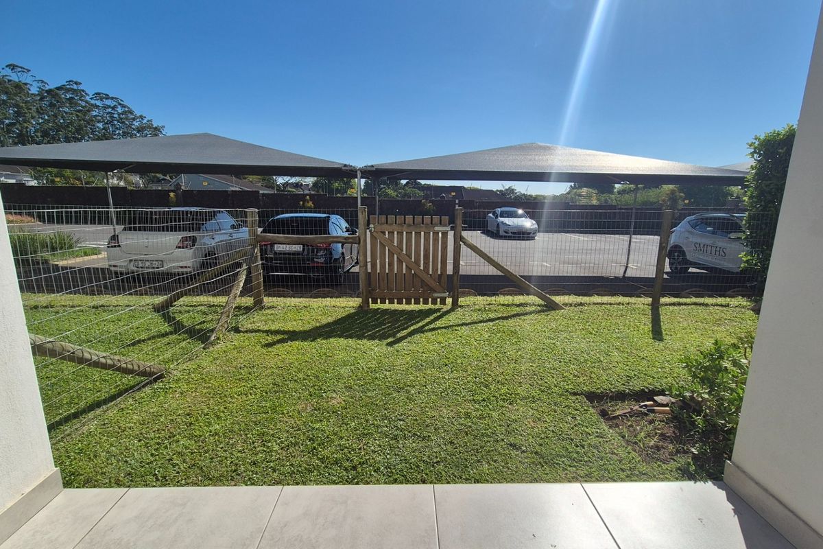 A view of a small private garden area featuring tiled patio flooring, wire mesh fencing with a wooden gate, and covered parking bays.