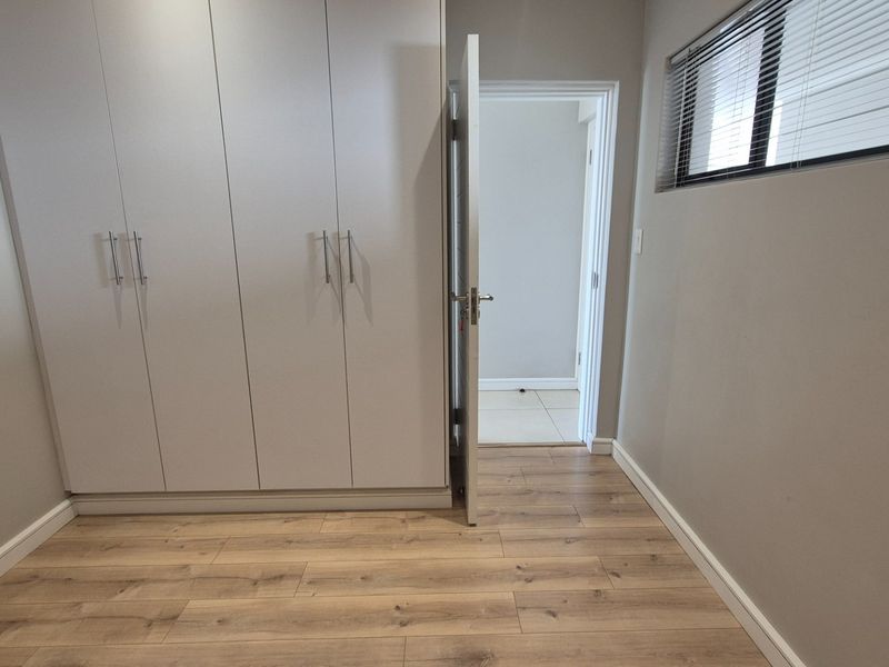 A clean, empty bedroom featuring modern light-toned laminate flooring, built-in cupboards, and a window fitted with horizontal blinds.