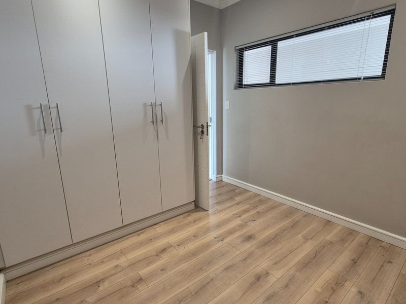 A clean, empty bedroom featuring modern built-in cupboards, light wood-look laminate flooring, and a high-set window with fitted blinds.