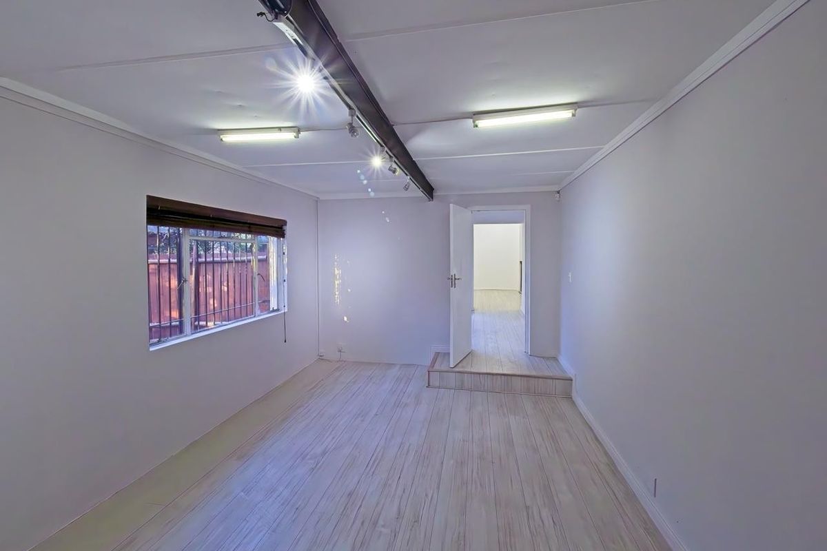 An empty, neutral-toned living area featuring laminate flooring, exposed ceiling beams, and fluorescent lighting, with a raised doorway leading to an adjacent room.