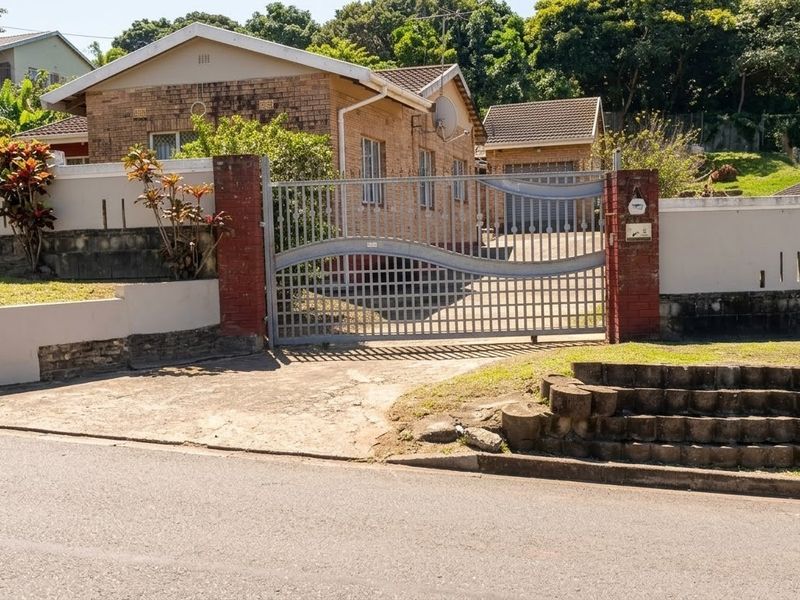 Front view of a brick house featuring a secure metal driveway gate, concrete driveway, and perimeter walling with established garden elements.