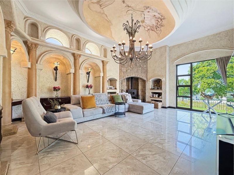 Spacious living room featuring a dramatic ceiling mural, stone fireplace, ornate wall niches with decorative columns, and polished marble flooring with garden access.