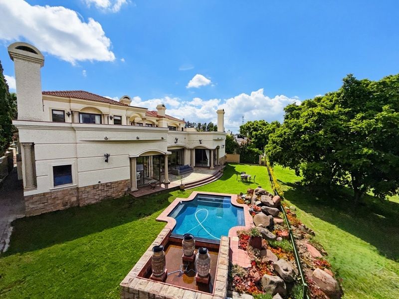 Spacious rear exterior featuring a well-maintained swimming pool, stone-accented water feature, covered patio, and expansive landscaped garden area.