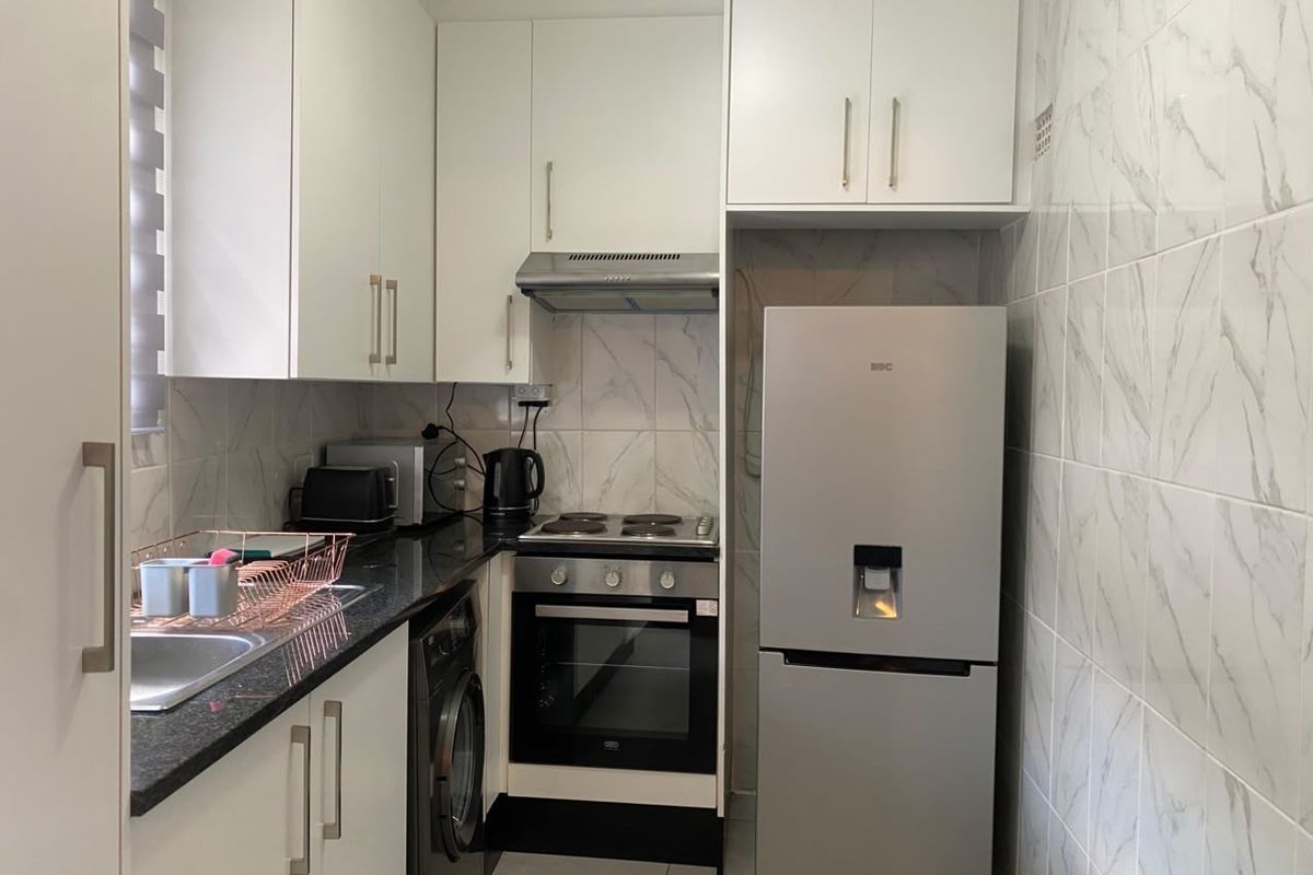 A compact, modern kitchen featuring white cabinetry, granite countertops, marble-look tiled walls, and integrated appliances including an oven and hob.