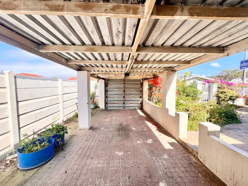 A covered carport area featuring a corrugated metal roof, timber support beams, and a paved driveway leading to a roll-up garage door.