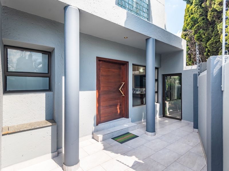 A modern front entrance featuring a solid timber door, tiled porch area, structural columns, and secure boundary wall with electric fencing.