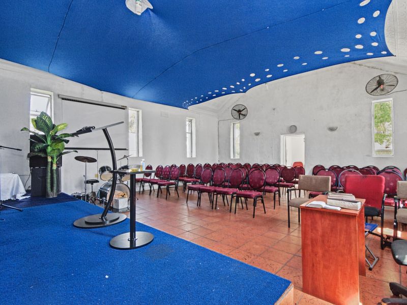 A multi-purpose hall featuring a raised stage, tiled flooring, and wall-mounted fans, suitable for community gatherings or events.