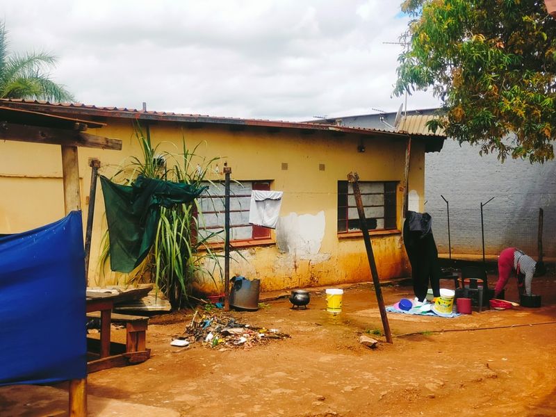 Rear exterior view of a property showing a dirt yard, weathered building facade with metal window frames, and security gate.