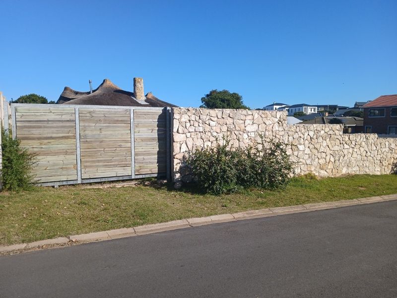 A street-level view of a residential property featuring a stone boundary wall and a timber gate, bordering a paved road.