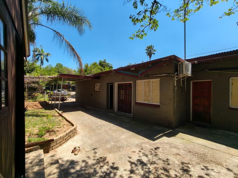 Rear exterior view of the property featuring a concrete driveway, covered carport area, and brick facade with established garden surroundings.