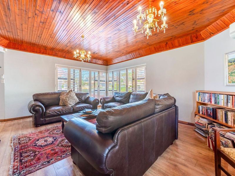 A spacious living room featuring a warm timber tongue and groove ceiling, laminate flooring, and plantation shutters, complemented by elegant chandeliers and air conditioning.