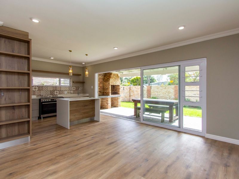 Spacious open-plan living area featuring a modern kitchen with island, laminate flooring, and large sliding doors opening to a covered patio with brick braai.