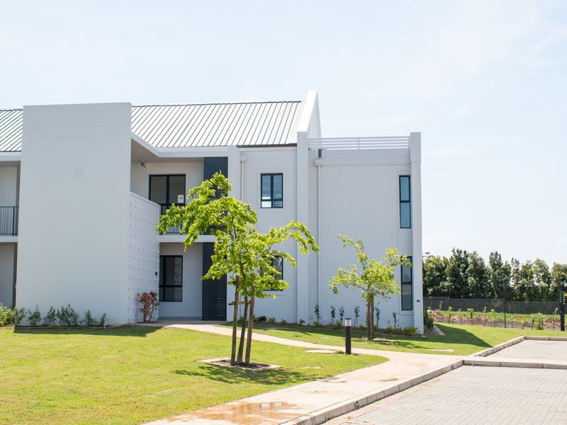A modern apartment building exterior featuring a clean white brick facade, metal roofing, and a well-maintained landscaped garden area with a paved walkway.