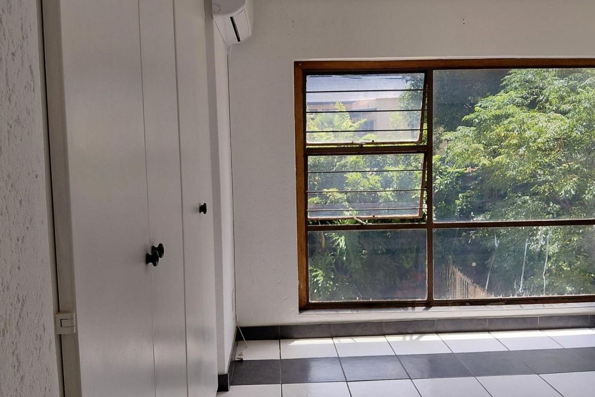 An empty bedroom featuring tiled flooring, built-in cupboards, and an air conditioning unit, with a large window overlooking greenery.