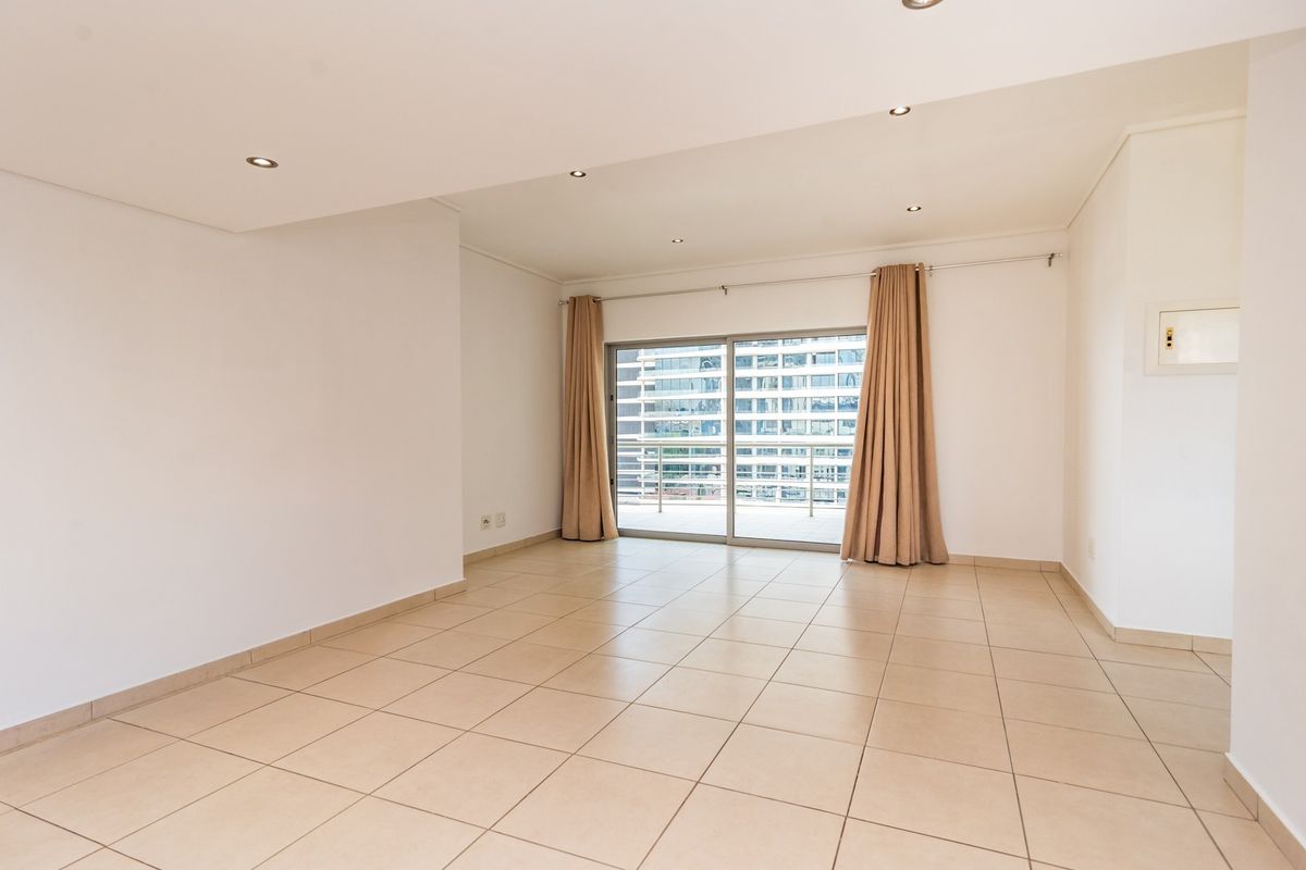 A bright, empty living room featuring neutral tiled flooring, recessed downlights, and a large sliding glass door leading to an outdoor balcony.