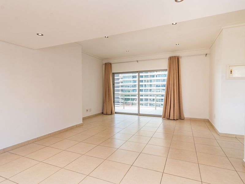 A bright, empty living room featuring neutral tiled flooring, recessed downlights, and a large sliding glass door leading to an outdoor balcony.